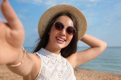 Photo of Beautiful young woman with straw hat and sunglasses taking selfie on beach. Stylish headdress