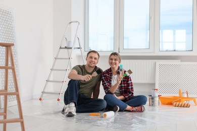 Photo of Happy couple with brushes and painting tools on floor in apartment during repair