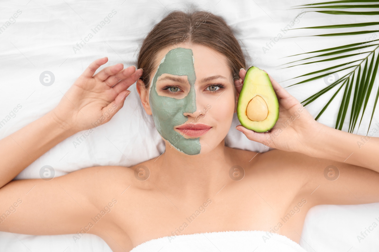 Photo of Beautiful woman holding avocado near her face with clay mask on white fabric, above view