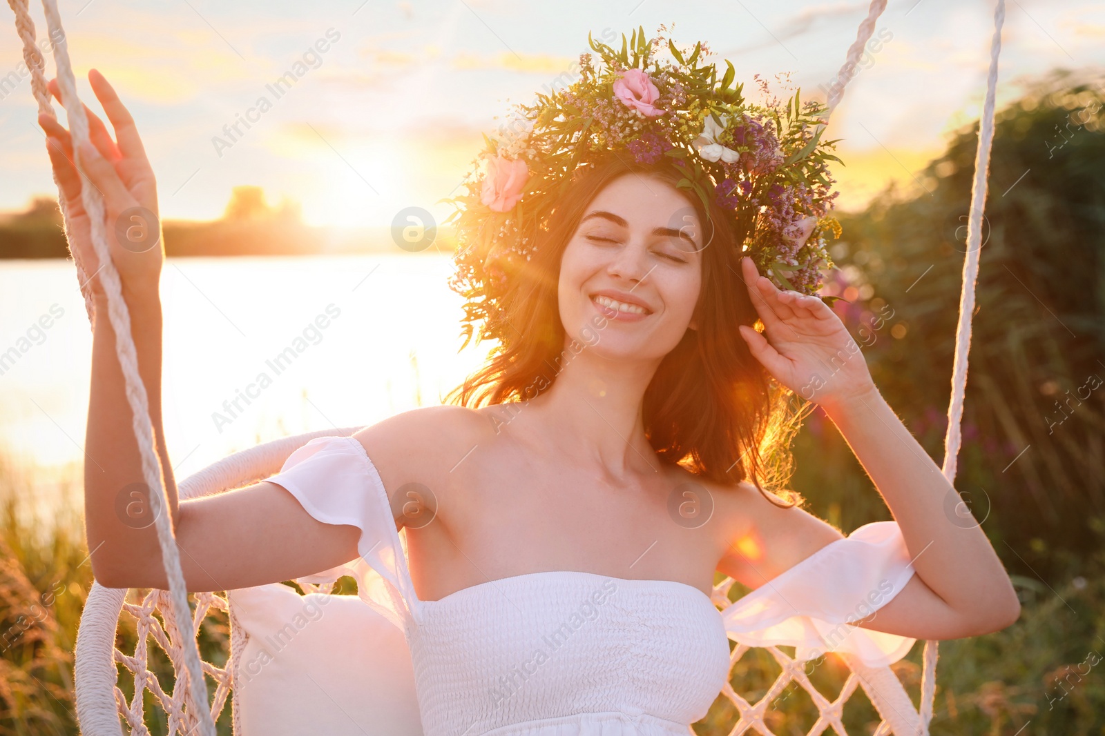 Photo of Young woman wearing wreath made of beautiful flowers on swing chair outdoors at sunset