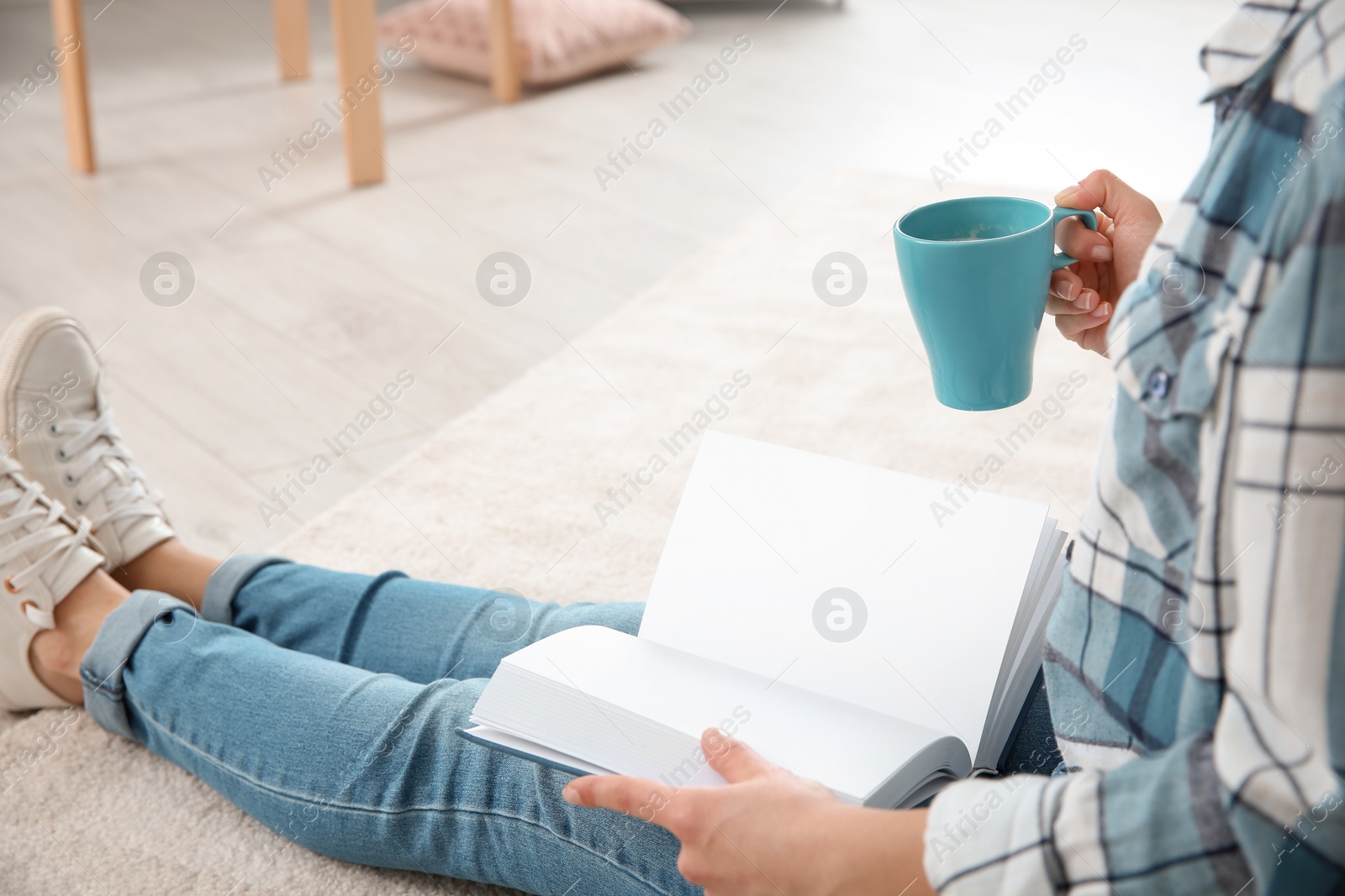 Photo of Beautiful young woman with cup of coffee reading book on floor at home