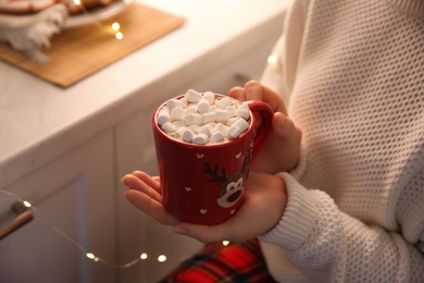 Photo of Woman holding cup of hot drink with marshmallow indoors, closeup