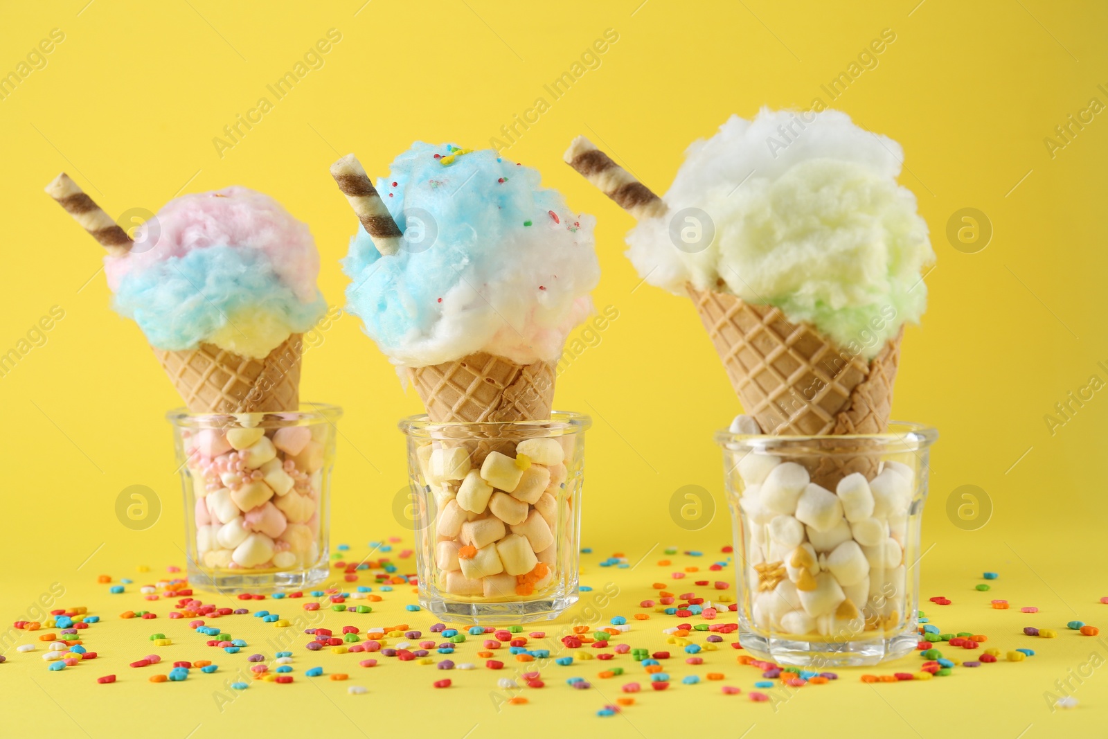 Photo of Sweet cotton candies in waffle cones and marshmallows on yellow background, closeup