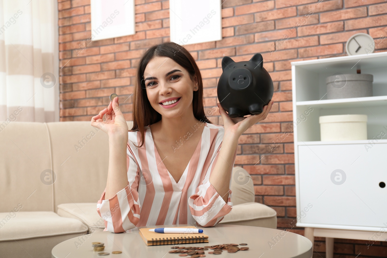 Photo of Happy young woman with piggy bank and money at home