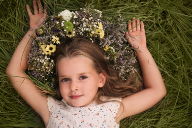 Photo of Cute little girl wearing wreath made of beautiful flowers on green grass, top view