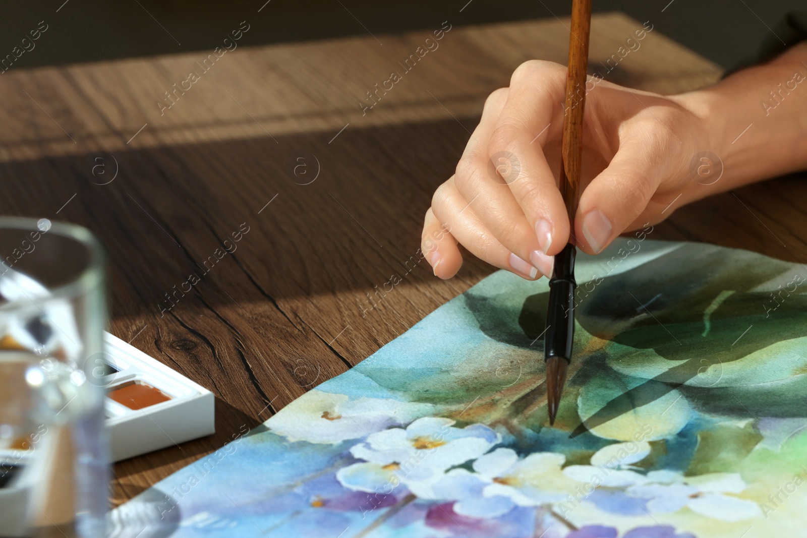 Photo of Woman painting flowers with watercolor at wooden table, closeup. Creative artwork