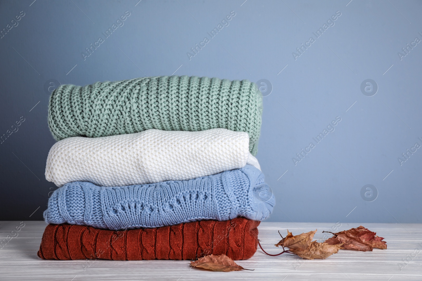 Photo of Stack of folded knitted sweaters and autumn leaves on white wooden table against grey background. Space for text