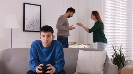 Photo of Teenage boy playing videogame while his parents arguing on background. Problems at home