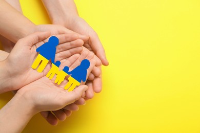 People holding paper family cutout in colors of Ukrainian flag on yellow background, top view. Space for text