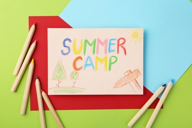 Photo of Paper with written text SUMMER CAMP, drawings and different pencils on color background, flat lay