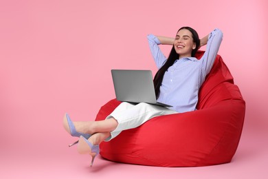 Happy woman with laptop sitting on beanbag chair against pink background