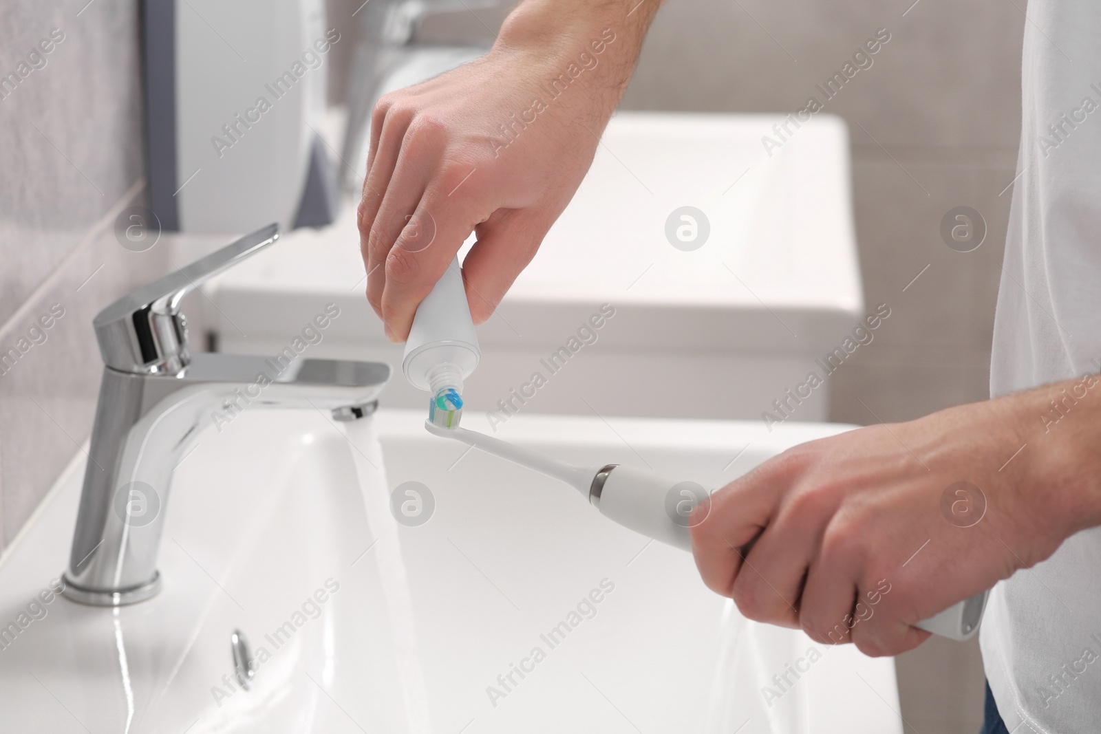 Photo of Man squeezing toothpaste from tube onto electric toothbrush above sink in bathroom, closeup