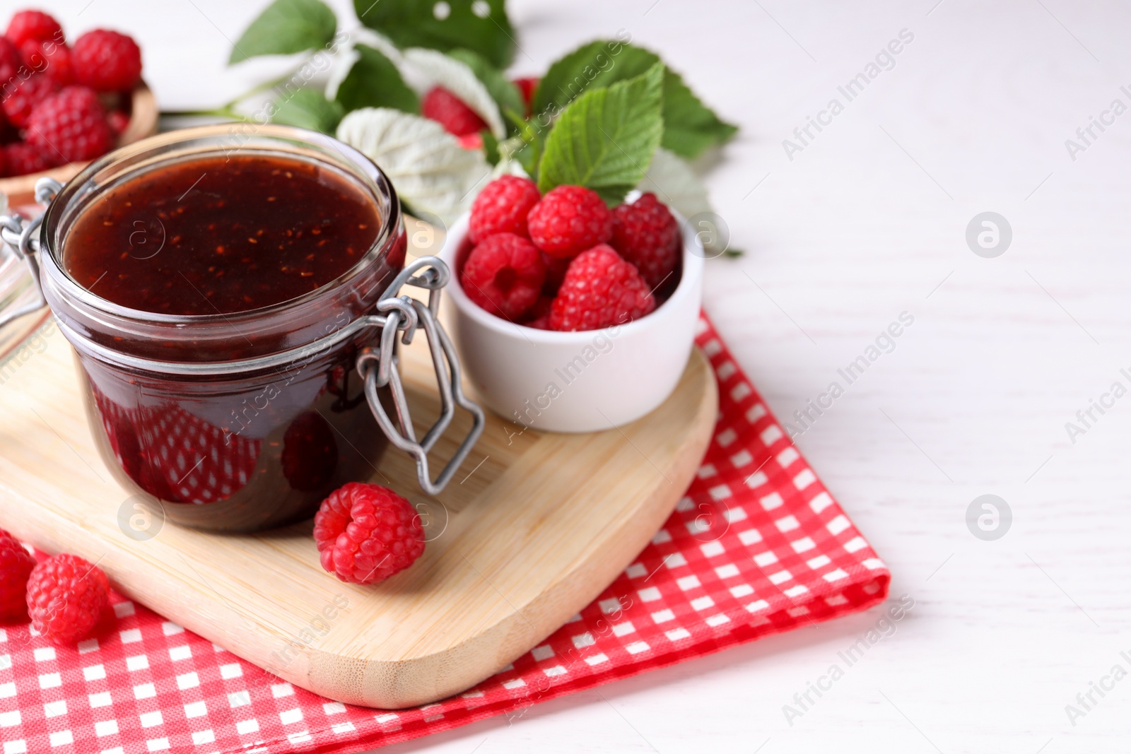 Photo of Jar of delicious raspberry jam, fresh berries and green leaves on white wooden table. Space for text