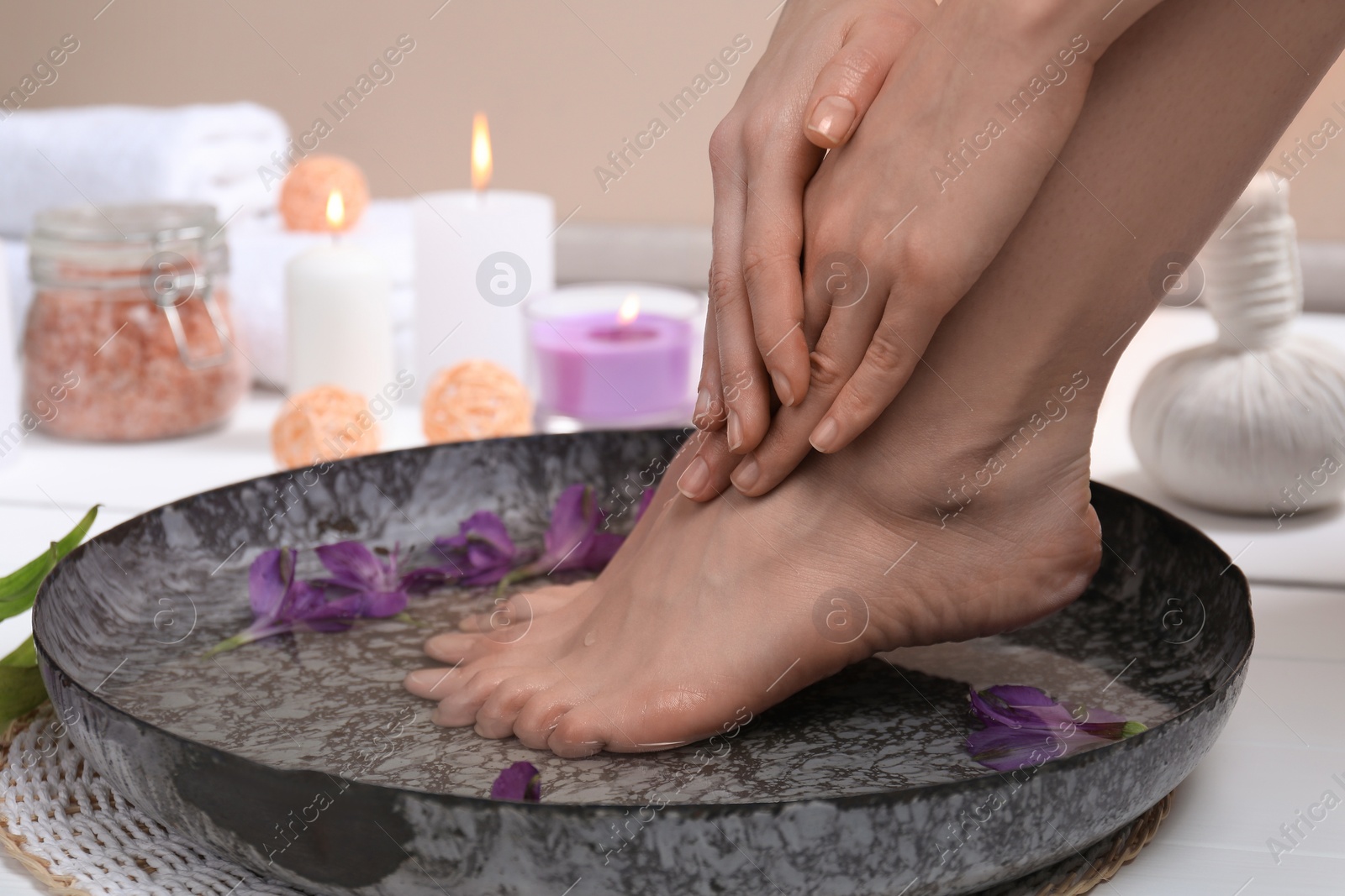 Photo of Woman soaking her feet in bowl with water and flowers on white floor, closeup. Spa treatment