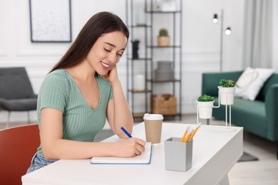 Young woman writing in notebook at white table indoors