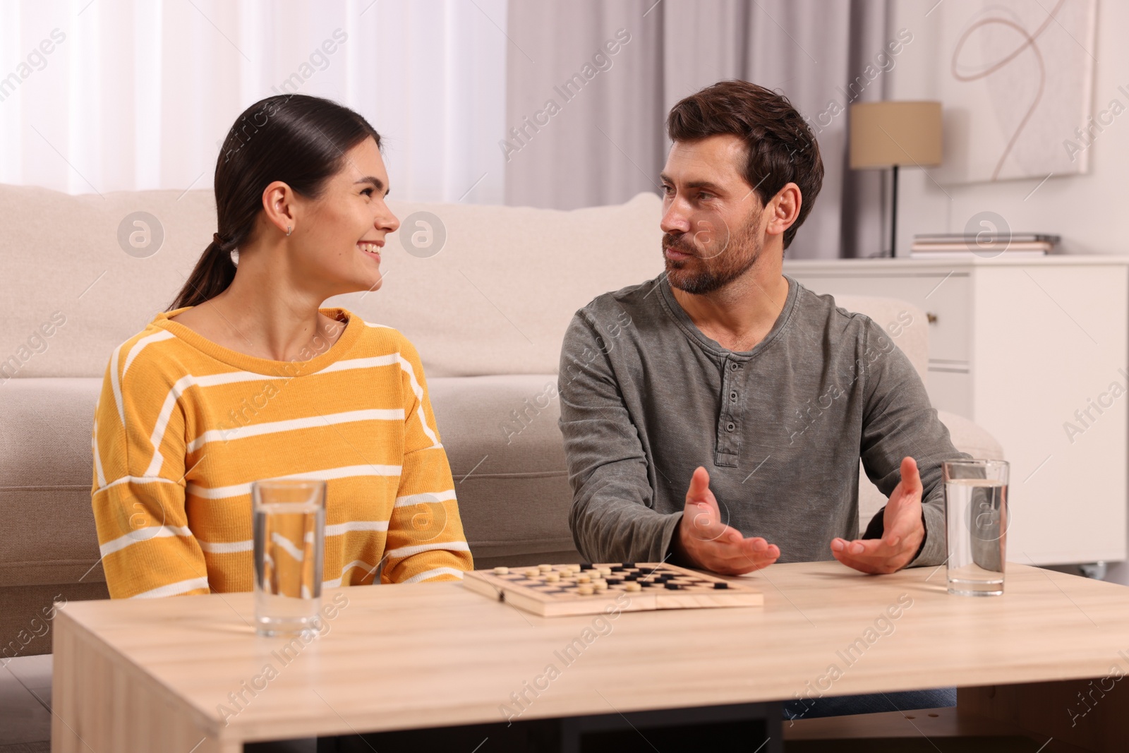 Photo of Happy couple talking while playing checkers at wooden table in room