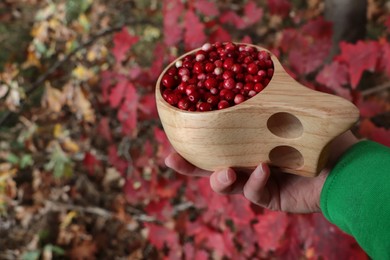 Photo of Woman holding tasty lingonberries in wooden cup near red leaves outdoors, closeup