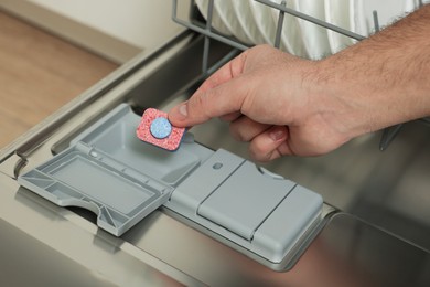 Photo of Man putting detergent tablet into open dishwasher indoors, closeup