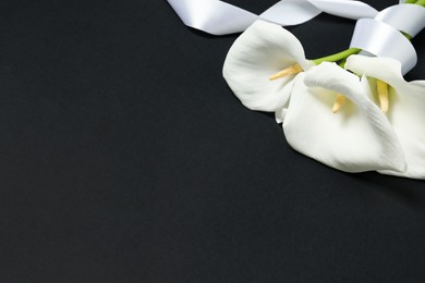 Photo of Beautiful calla lilies and white ribbon on black background, closeup with space for text. Funeral symbol