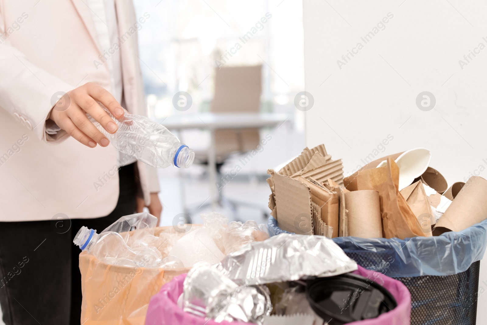 Photo of Woman putting used plastic bottle into trash bin in office, closeup. Waste recycling