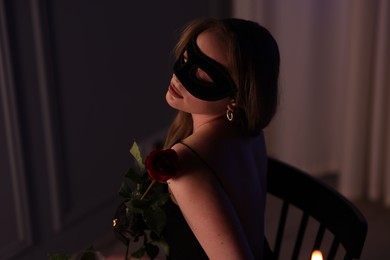Photo of Elegant woman in black eye mask with rose indoors in evening