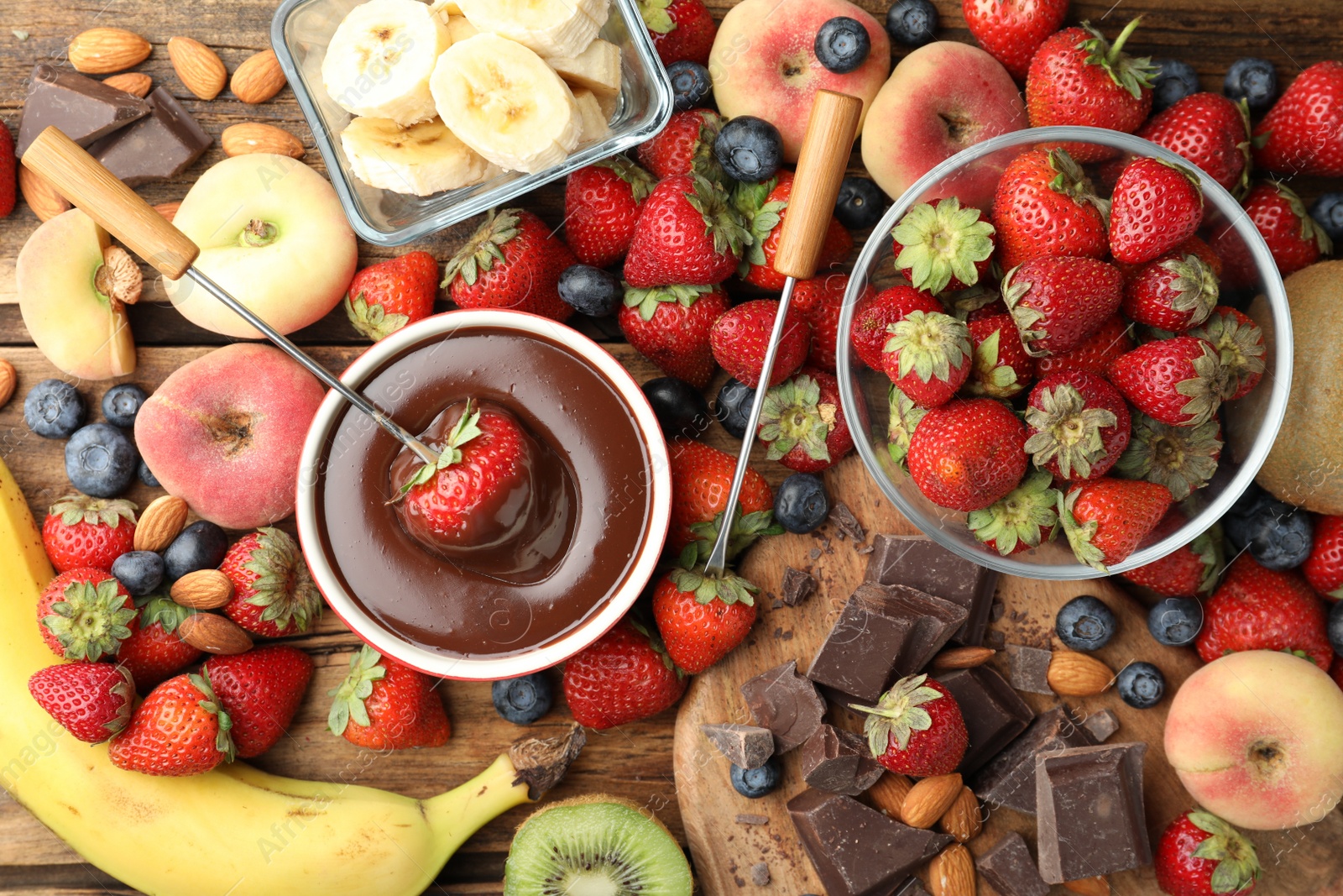 Photo of Fondue fork with strawberry in bowl of melted chocolate surrounded by other fruits on wooden table, flat lay