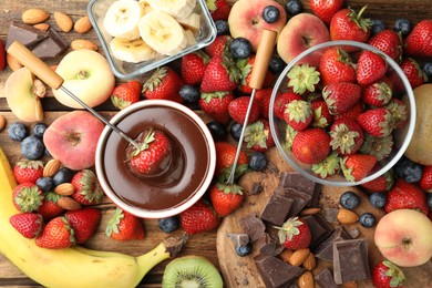 Photo of Fondue fork with strawberry in bowl of melted chocolate surrounded by other fruits on wooden table, flat lay