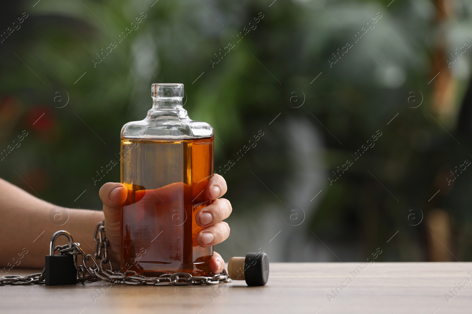 Photo of Addicted man chained to bottle of alcoholic drink at table against blurred background, closeup. Space for text