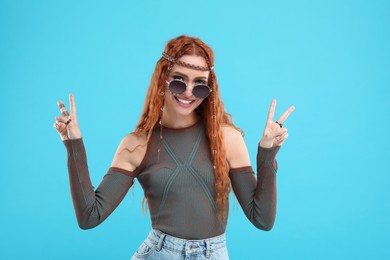 Photo of Stylish young hippie woman in sunglasses showing V-sign on light blue background