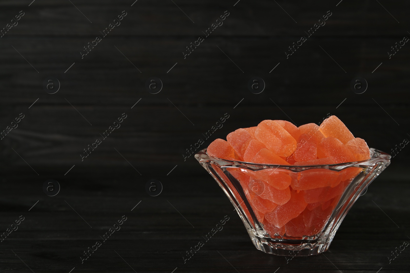 Photo of Tasty orange jelly candies in glass bowl on black wooden table. Space for text
