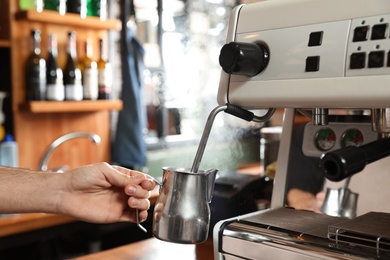 Photo of Barista steaming milk in metal jug with coffee machine wand at bar counter, closeup