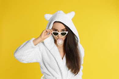 Photo of Young woman in bathrobe and sunglasses blowing chewing gum on yellow background