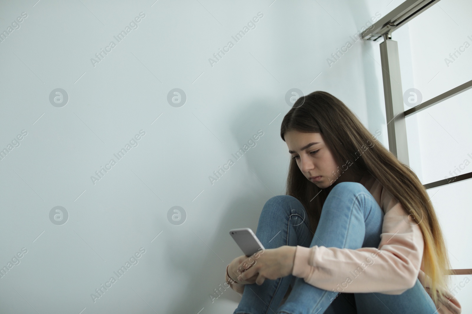 Photo of Upset teenage girl with smartphone sitting at wall indoors. Space for text