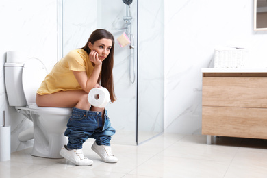 Photo of Upset woman with paper roll sitting on toilet bowl in bathroom
