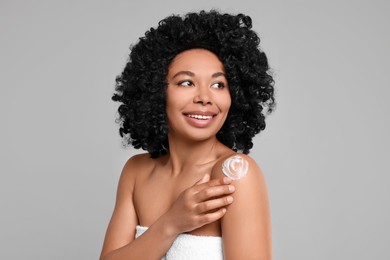 Young woman applying body cream onto shoulder on grey background