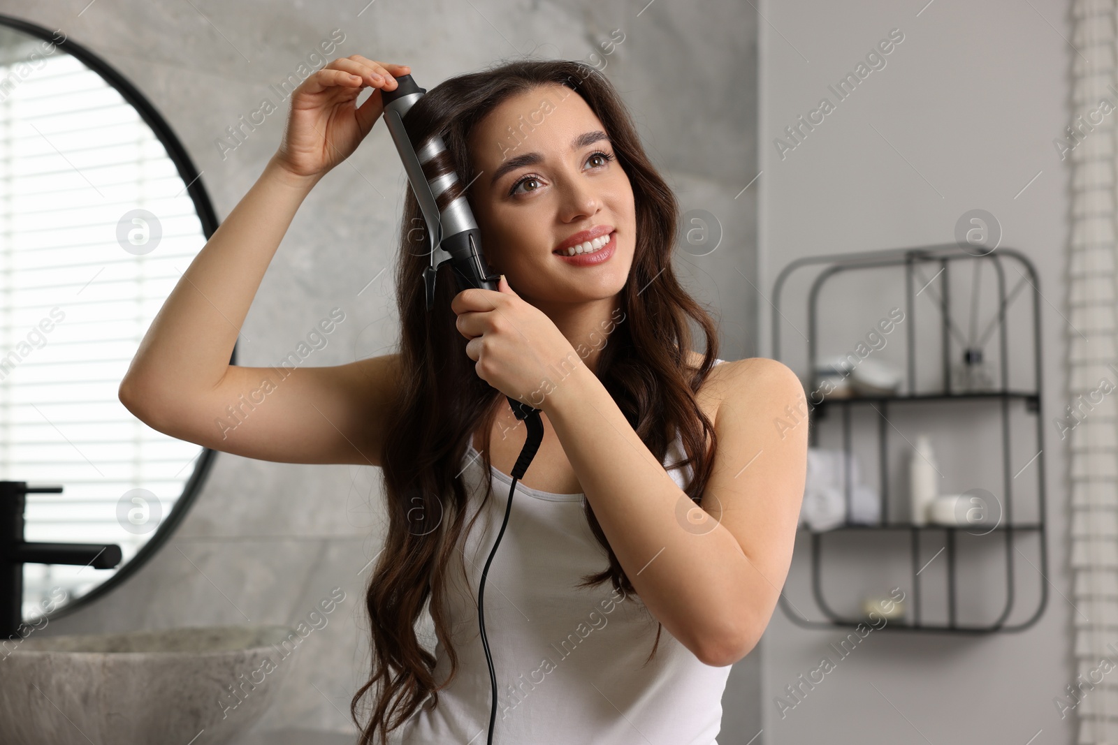 Photo of Smiling woman using curling hair iron in bathroom