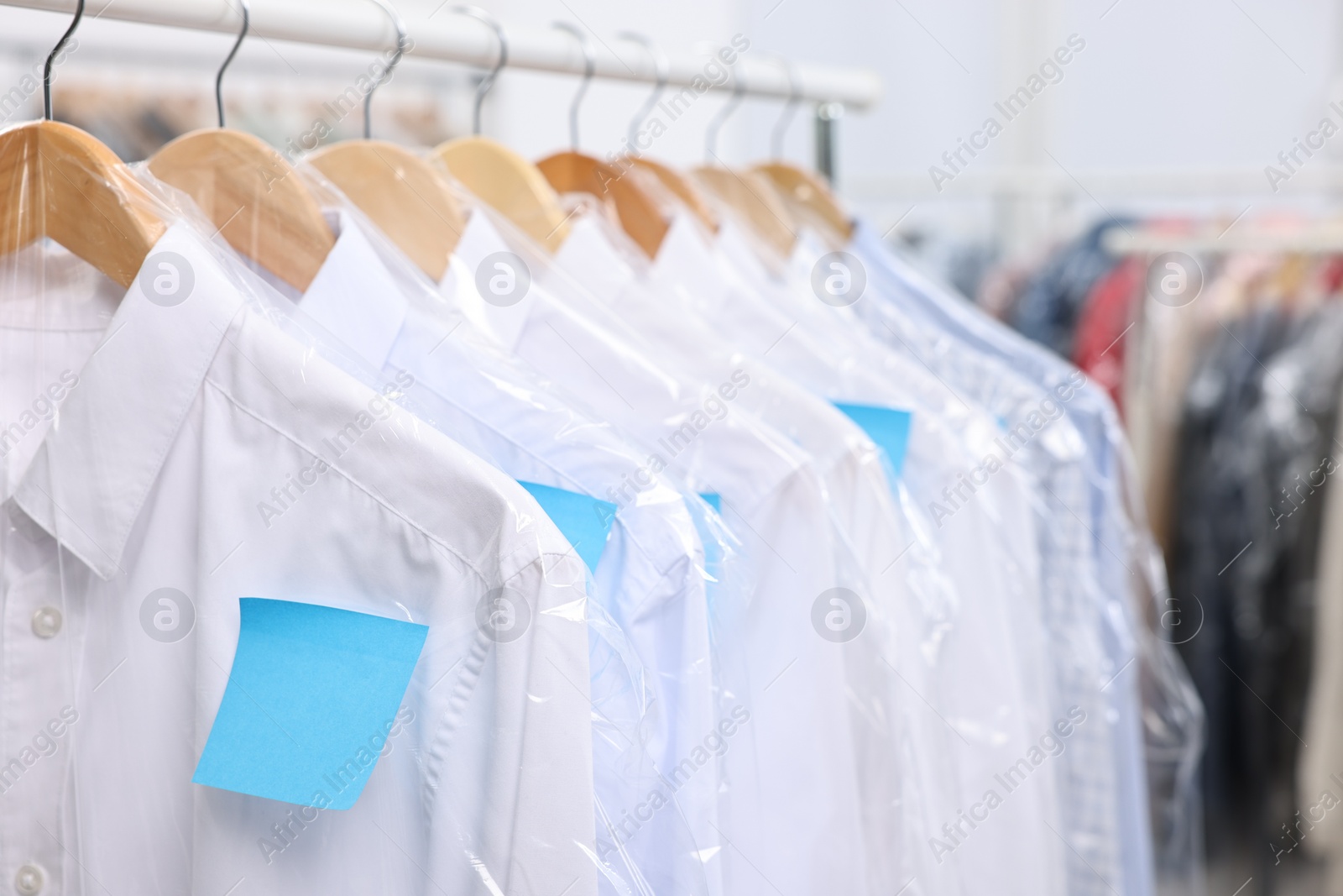 Photo of Dry-cleaning service. Many different clothes in plastic bags hanging on rack indoors, closeup