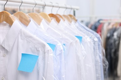 Photo of Dry-cleaning service. Many different clothes in plastic bags hanging on rack indoors, closeup