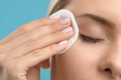 Woman removing makeup with cotton pad on light blue background, closeup