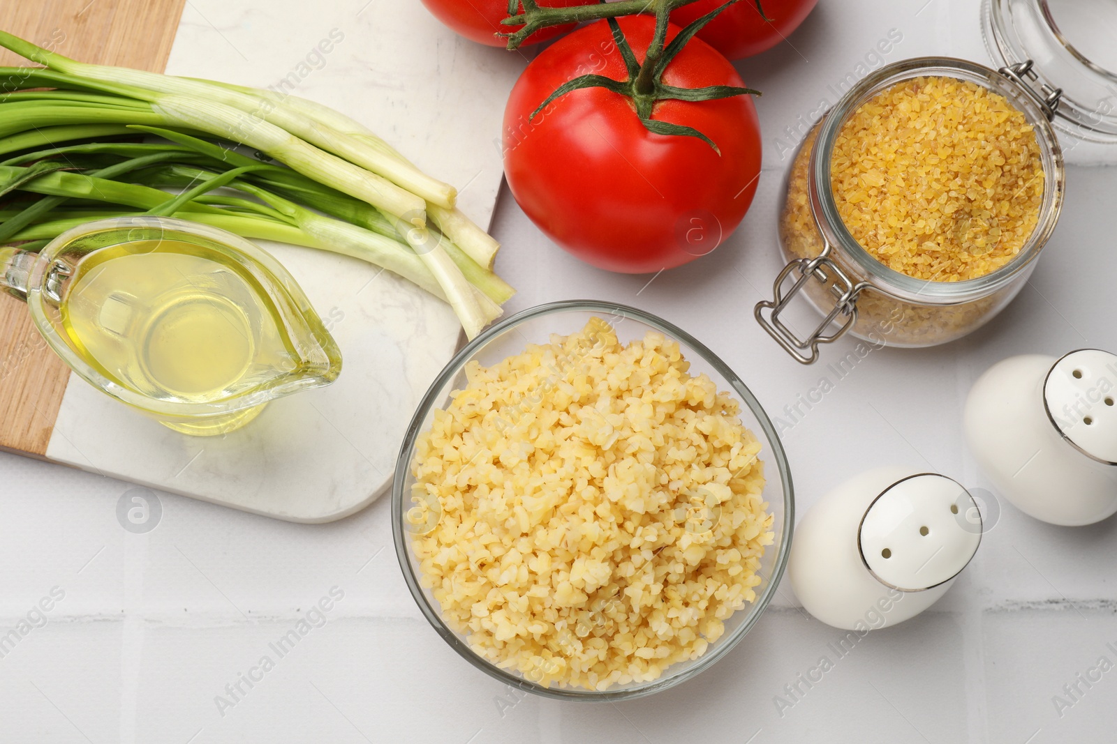 Photo of Delicious bulgur in bowl, vegetables, oil and spices on white tiled table, flat lay