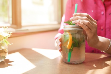 Photo of Young woman with mason jar of tasty natural lemonade in cafe, closeup. Detox drink