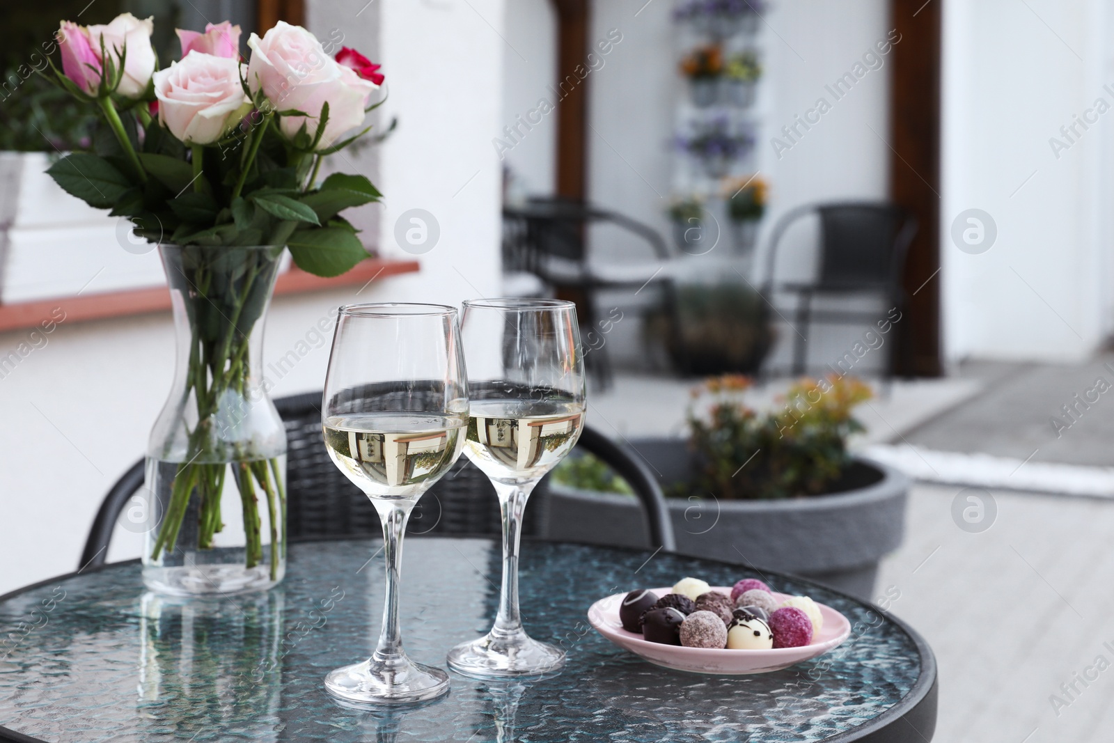 Photo of Vase with roses, glasses of wine and candies on glass table near house on outdoor terrace
