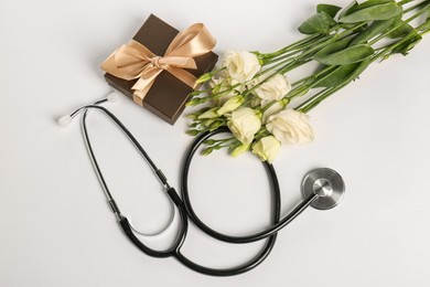 Photo of Stethoscope, gift box and eustoma flowers on white background, flat lay. Happy Doctor's Day