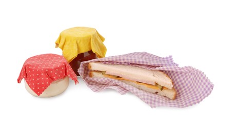 Photo of Tasty sandwich and jars of sauces in beeswax food wraps on white background