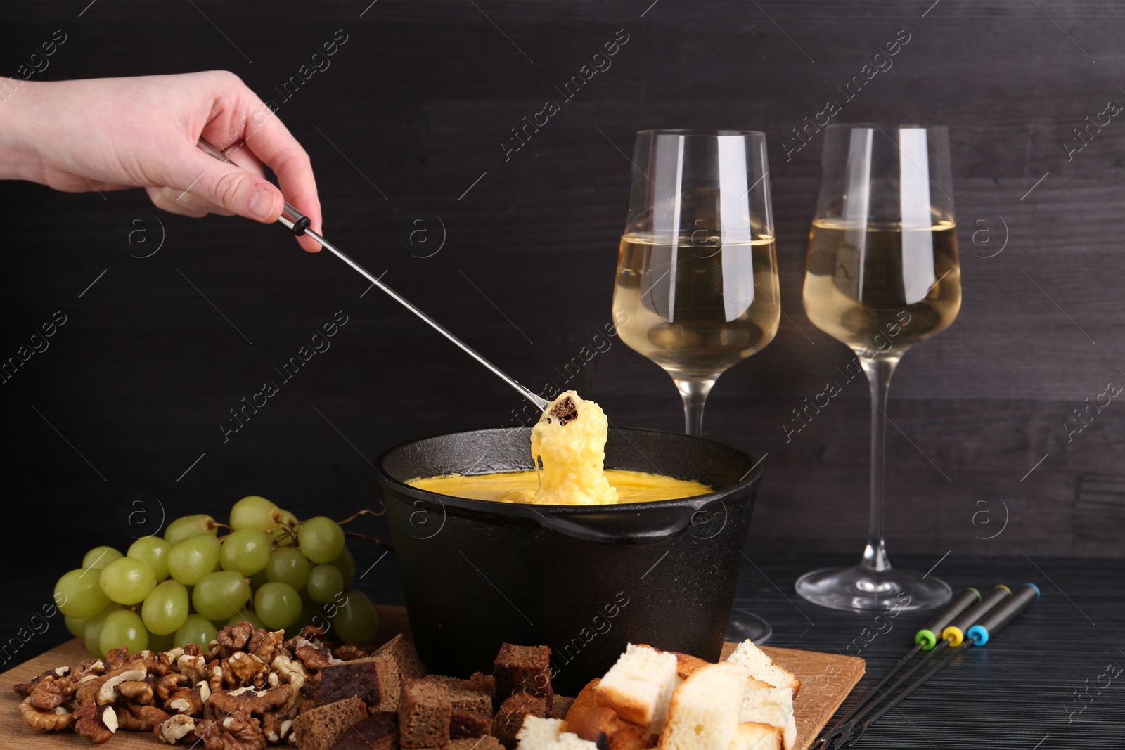 Photo of Woman dipping piece of bread into fondue pot with melted cheese at black wooden table, closeup