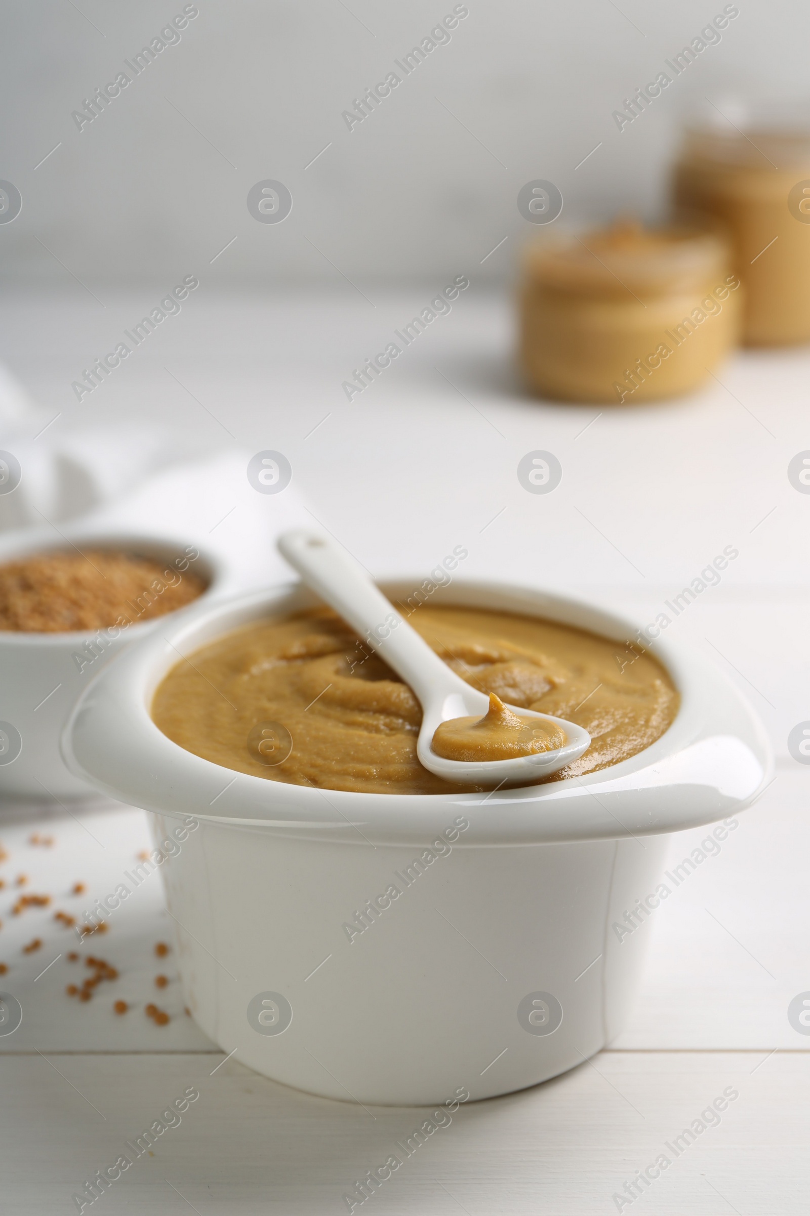 Photo of Spoon and bowl of tasty mustard sauce on white wooden table, closeup