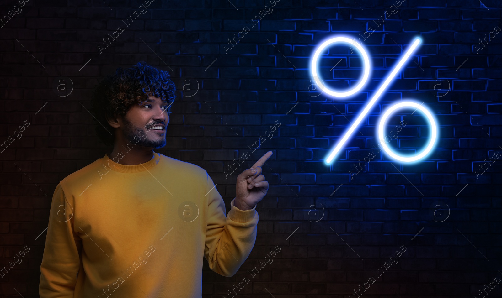 Image of Discount offer. Happy man pointing at neon percent sign on brick wall in darkness, banner design