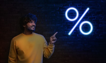 Image of Discount offer. Happy man pointing at neon percent sign on brick wall in darkness, banner design