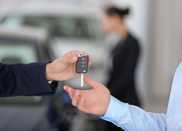 Young salesman giving car key to client against blurred background, closeup