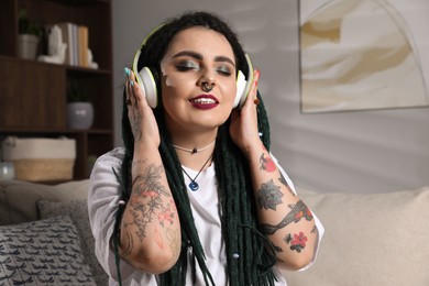 Photo of Beautiful young woman with tattoos on body, nose piercing and dreadlocks listening to music at home